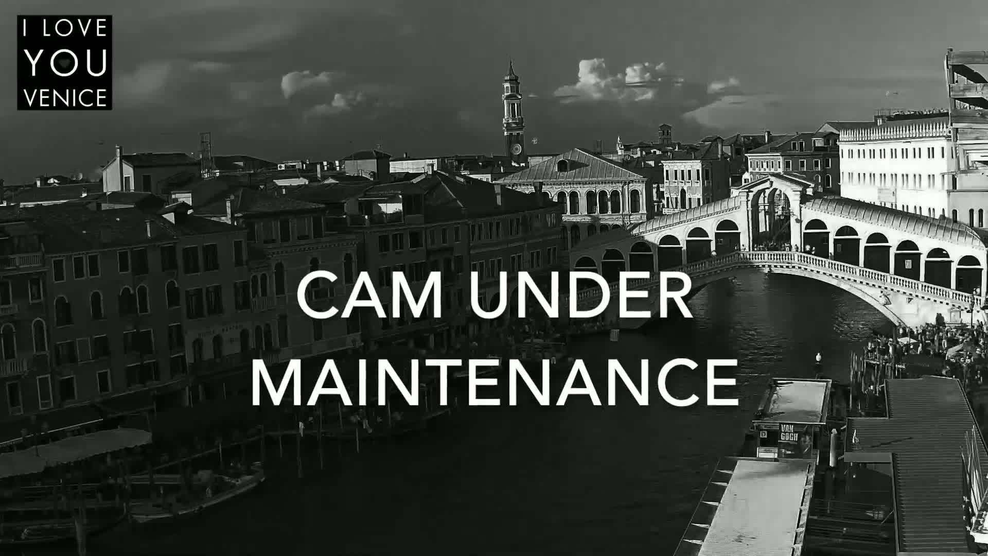 A black and white, high-angle view shows a Venetian bridge over a canal, with buildings lining the waterway under a partly cloudy sky.