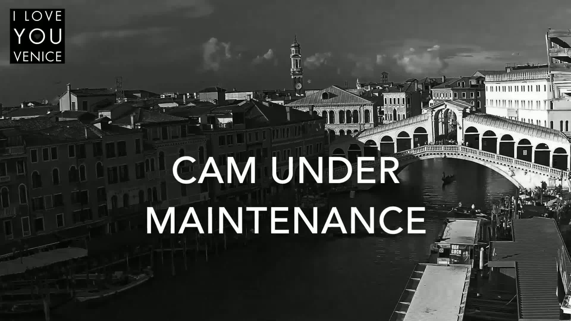 A black and white aerial view shows a canal in Venice, Italy, with a bridge and buildings under a partly cloudy sky.