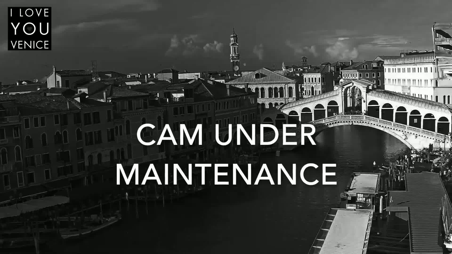 A black and white, high-angle view of a canal in Venice, Italy, showing a bridge and buildings under a partly cloudy sky.