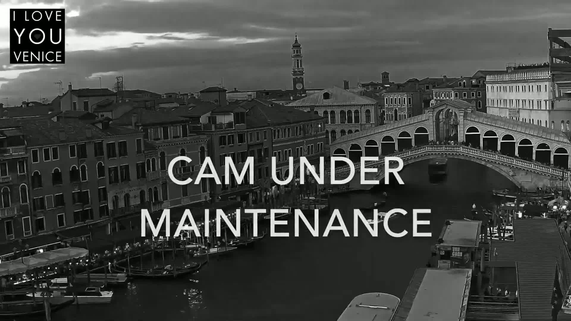 A black and white, high-angle view of Venice, Italy at dusk, showing a canal with gondolas and boats, a bridge, and numerous buildings.