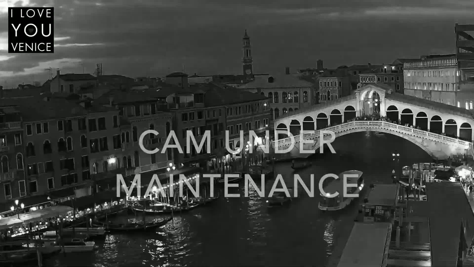 A nighttime, black and white view of a canal in Venice, with boats moored along the sides and a bridge spanning the waterway.