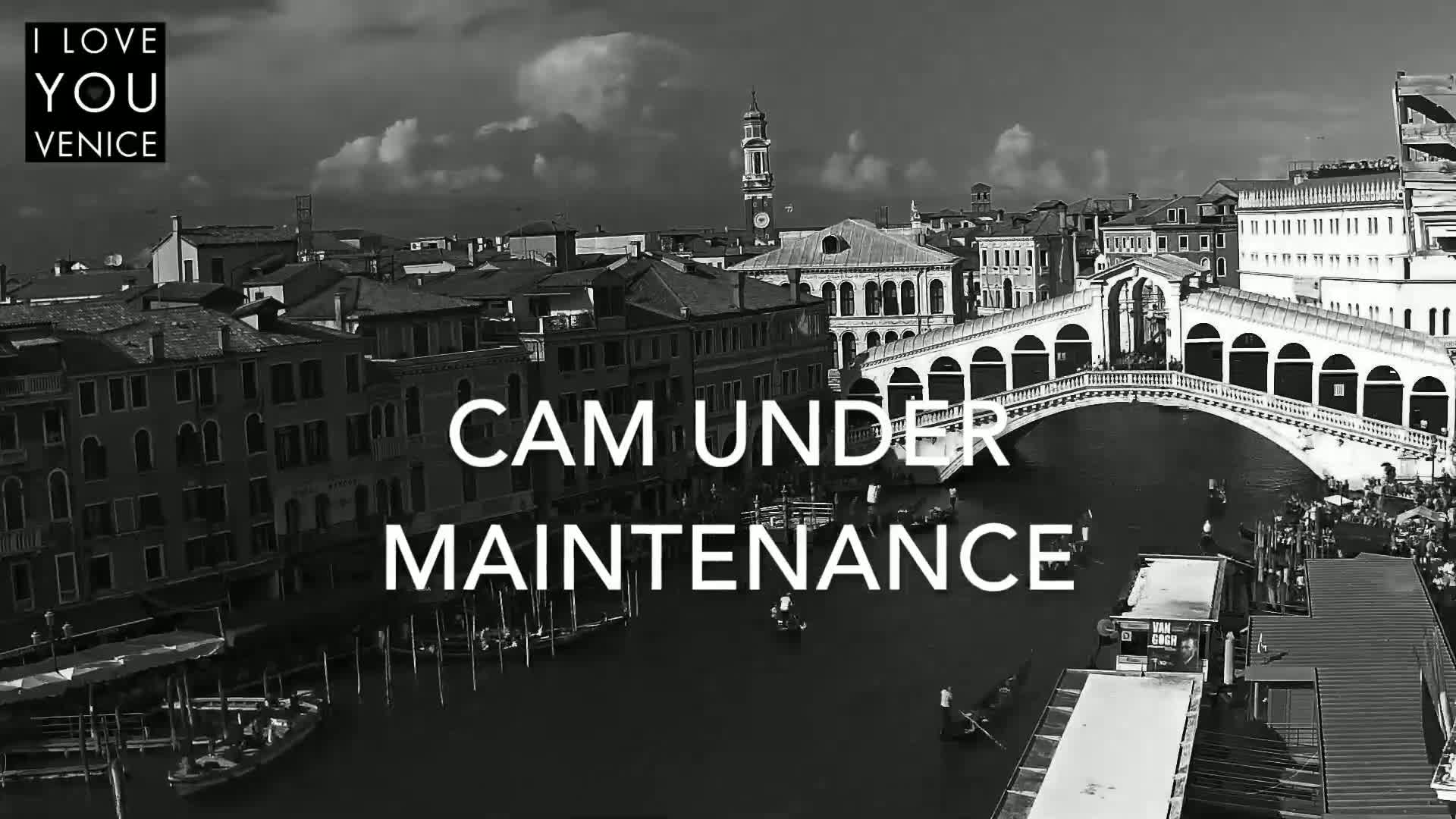 A black and white, high-angle view shows a canal in Venice, Italy, with gondolas and a bridge, under a partly cloudy sky.