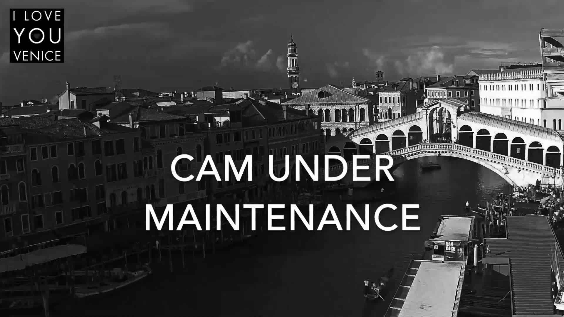 A black and white aerial view captures the historic Rialto Bridge spanning a canal bustling with boats, surrounded by classic Venetian buildings and a distant bell tower under a partly cloudy sky.