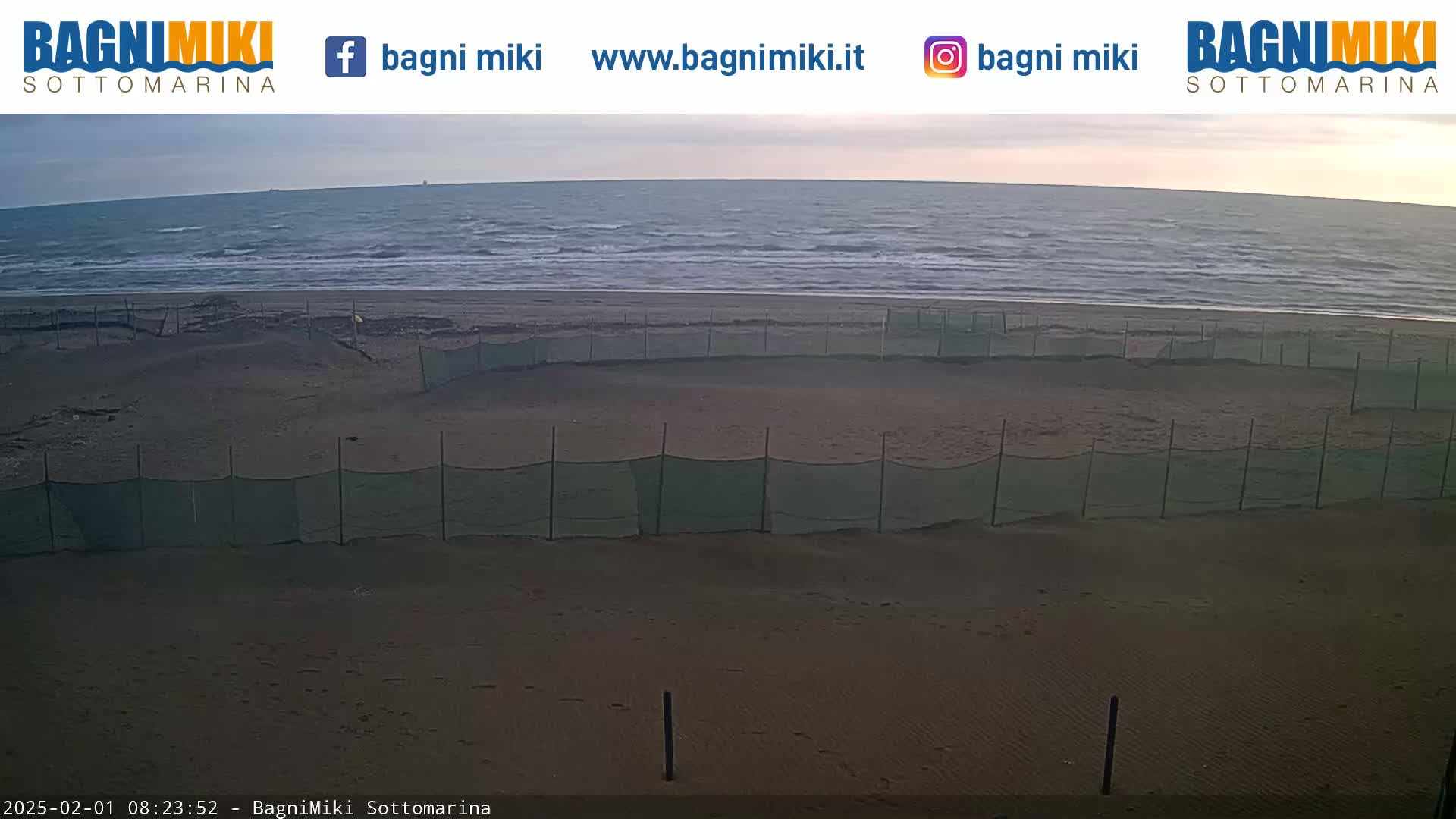 A sandy beach with several low green mesh fences stretches from a calm, gray-blue sea under a mostly cloudy sky.