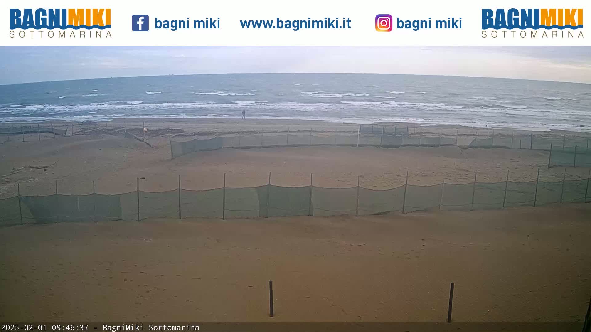 A sandy beach with a wavy sea under an overcast sky is partially enclosed by netting.