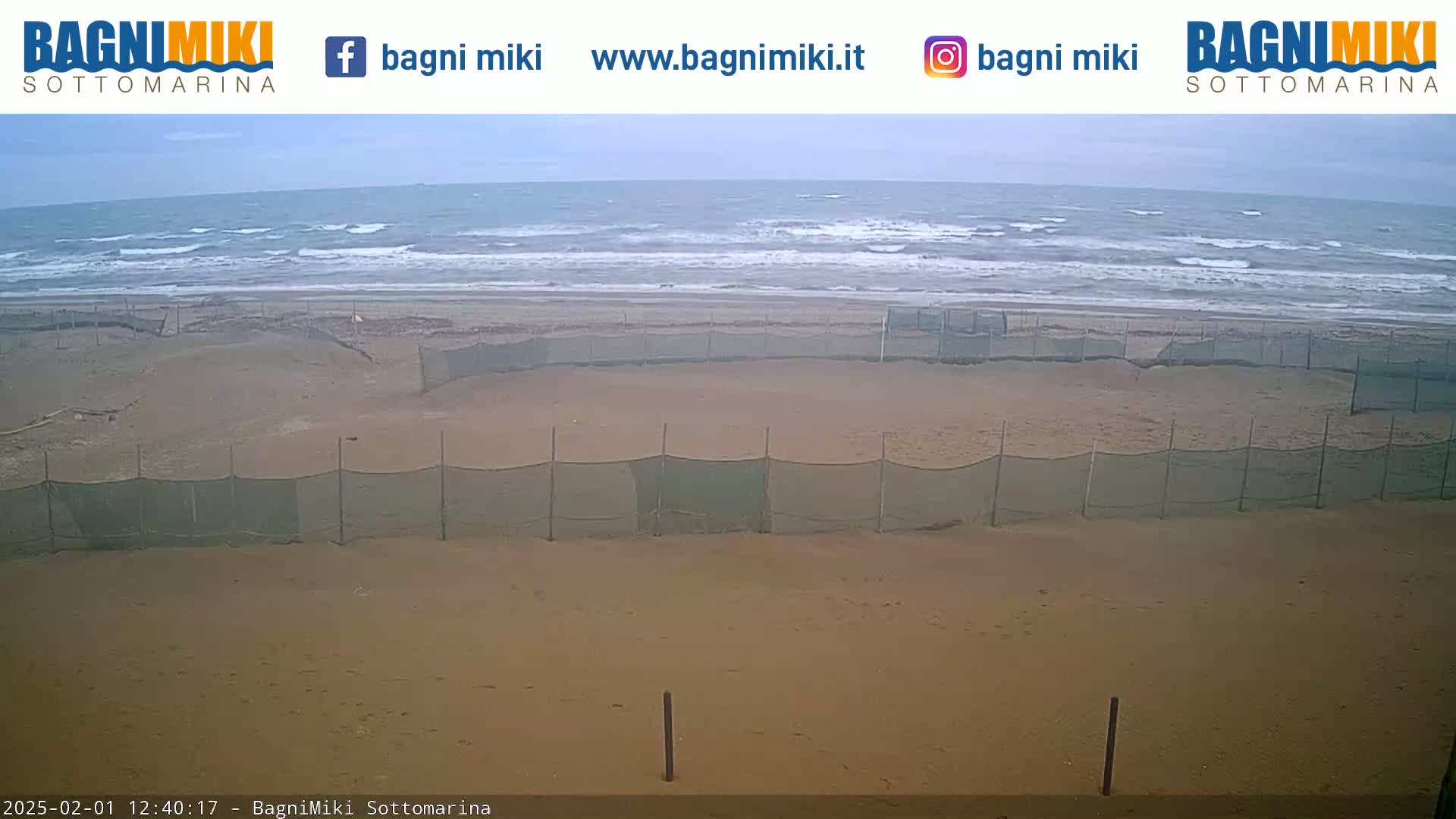 A sandy beach with several enclosed areas protected by netting is shown, with a somewhat choppy sea under an overcast sky in the background.