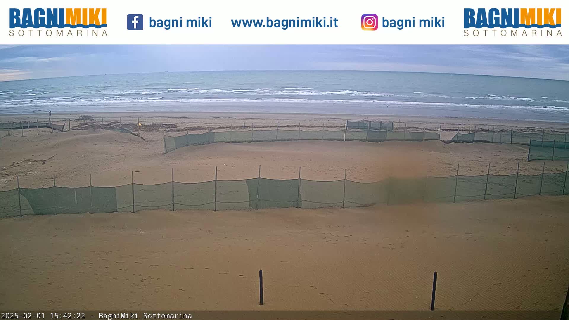 A sandy beach with several enclosed areas sectioned off by netting is visible, under a mostly cloudy sky with a calm ocean in the background.