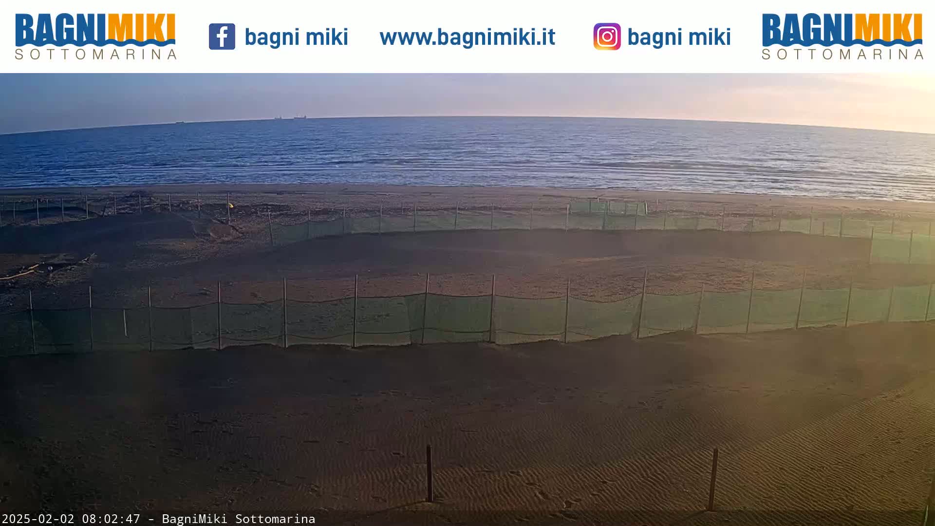 A dark sandy beach with several sections enclosed by netting is visible, overlooking a calm sea under a clear, sunrise sky.