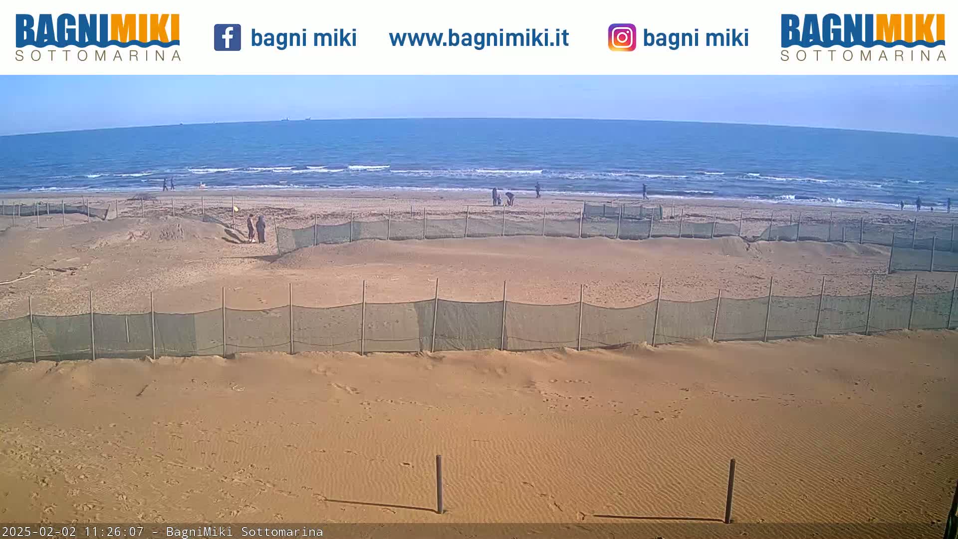 A sandy beach with a calm ocean and several people are visible in the background, with protective netting enclosing sections of the beach in the foreground under a clear sky.