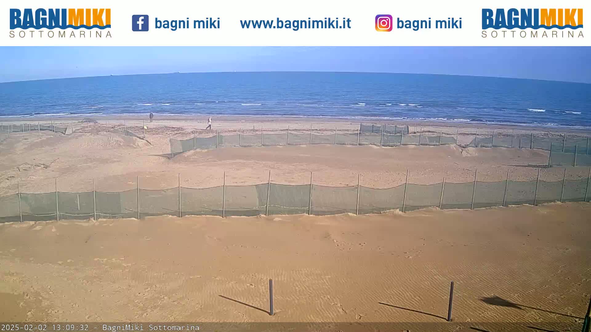 A sandy beach with several mesh fences is visible under a sunny sky, and a calm sea stretches beyond it.