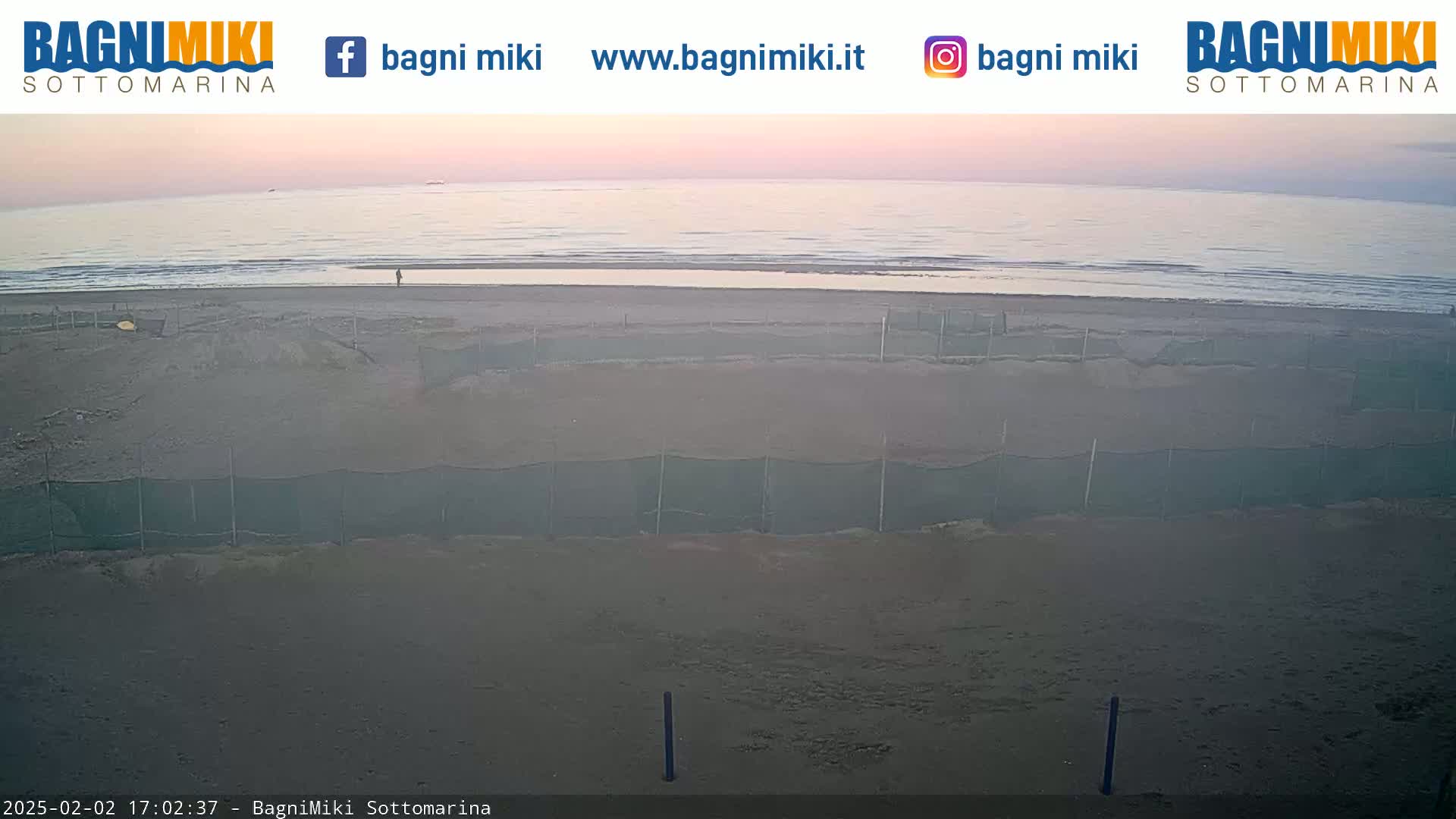 A calm, pale-pink sunrise illuminates a sandy beach with low-lying nets and a few people visible in the distance.
