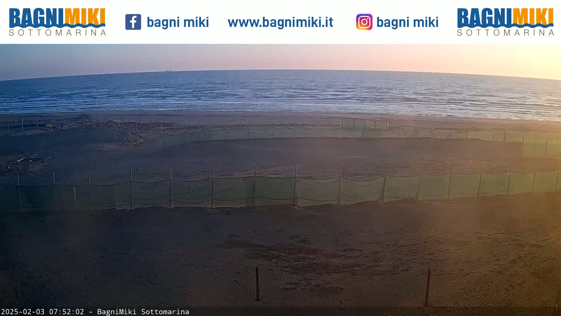 A sandy beach with several sections enclosed by netting is visible, overlooking a calm sea under a clear, sunrise sky.