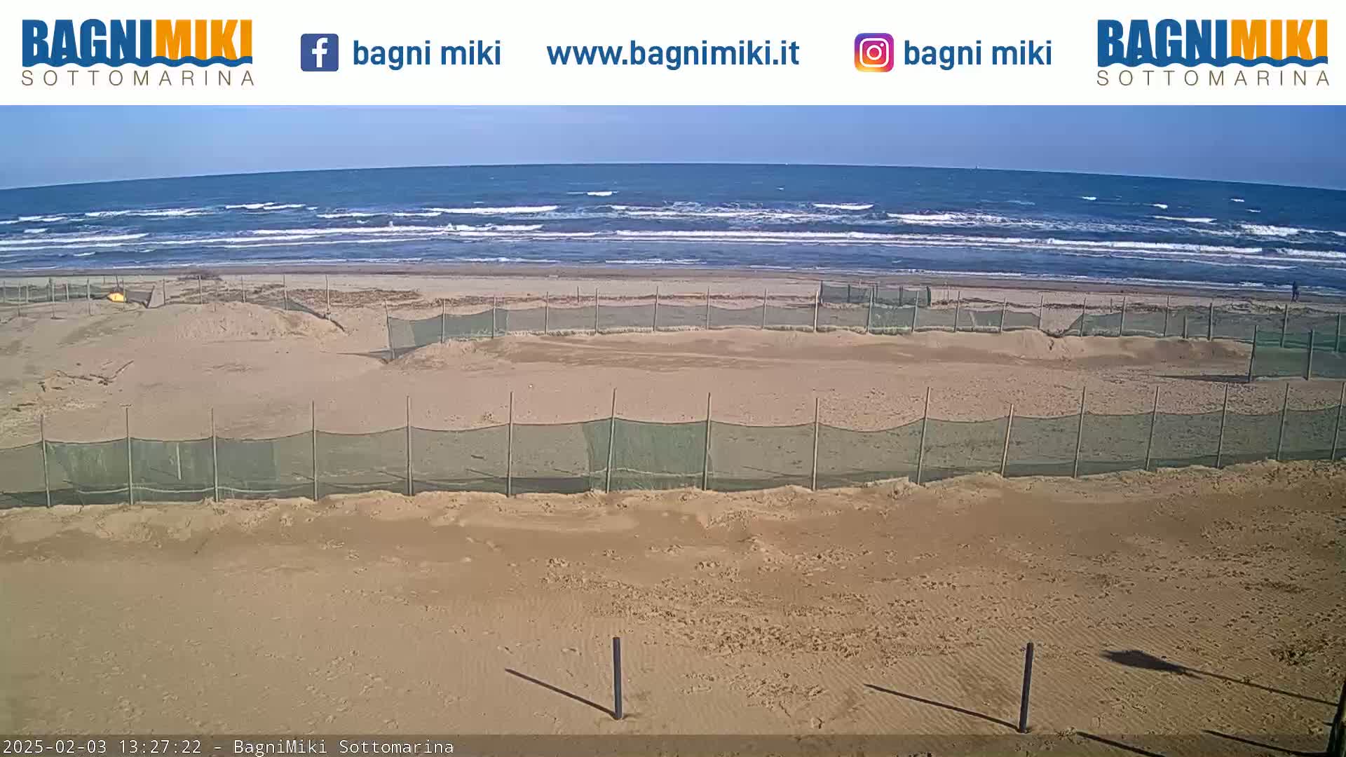 A sandy beach with gently rolling waves under a clear sky is sectioned off by several mesh fences.