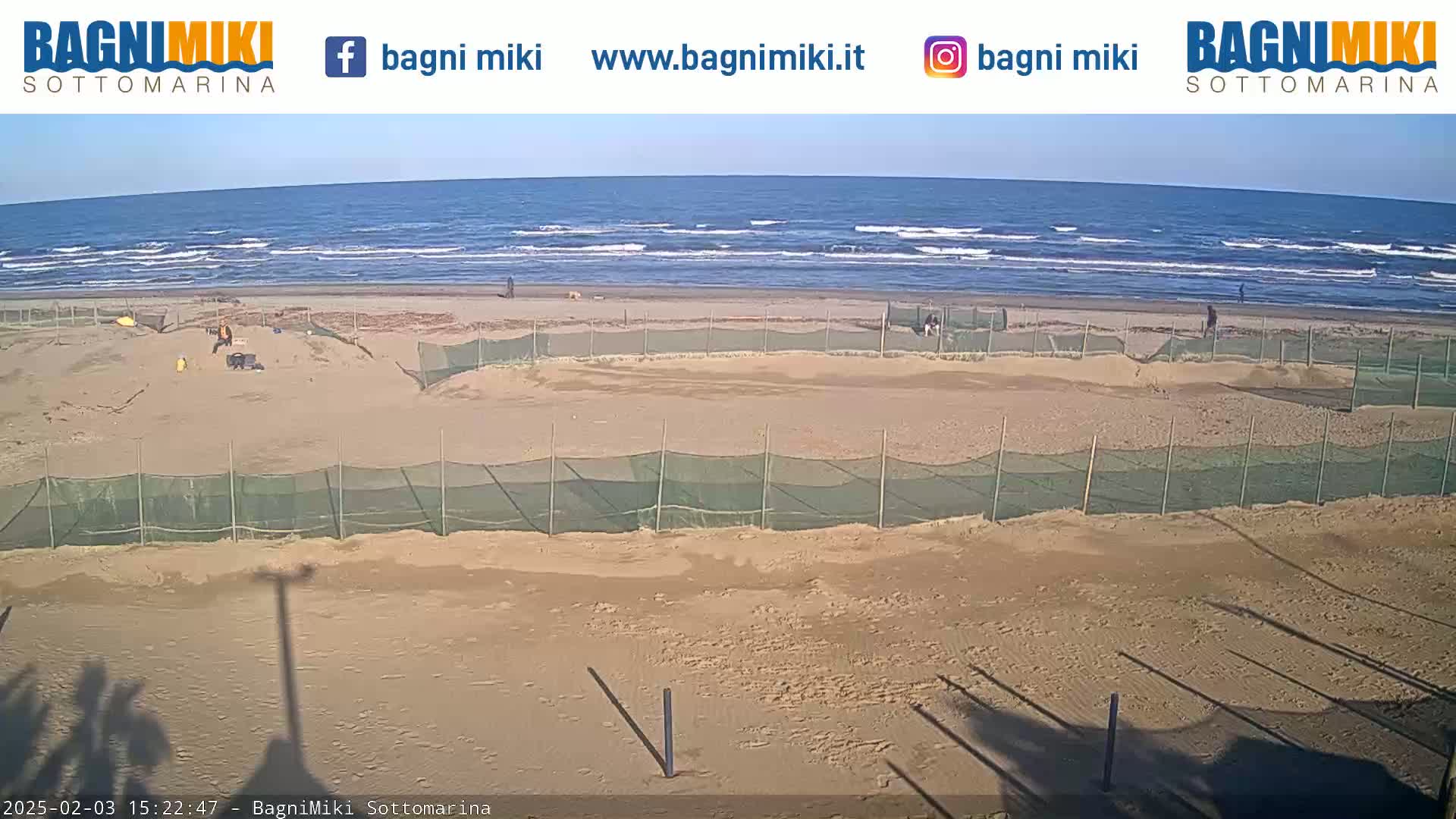 A sandy beach with several enclosed areas and people working near them is visible under a clear sky with a calm sea in the background.
