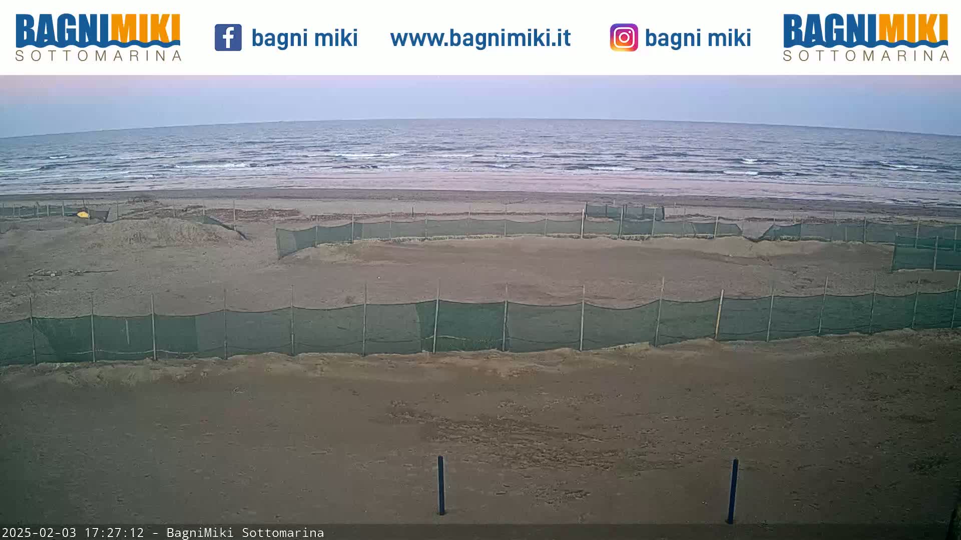 A sandy beach with several enclosed areas protected by netting is shown, overlooking a calm, overcast ocean.