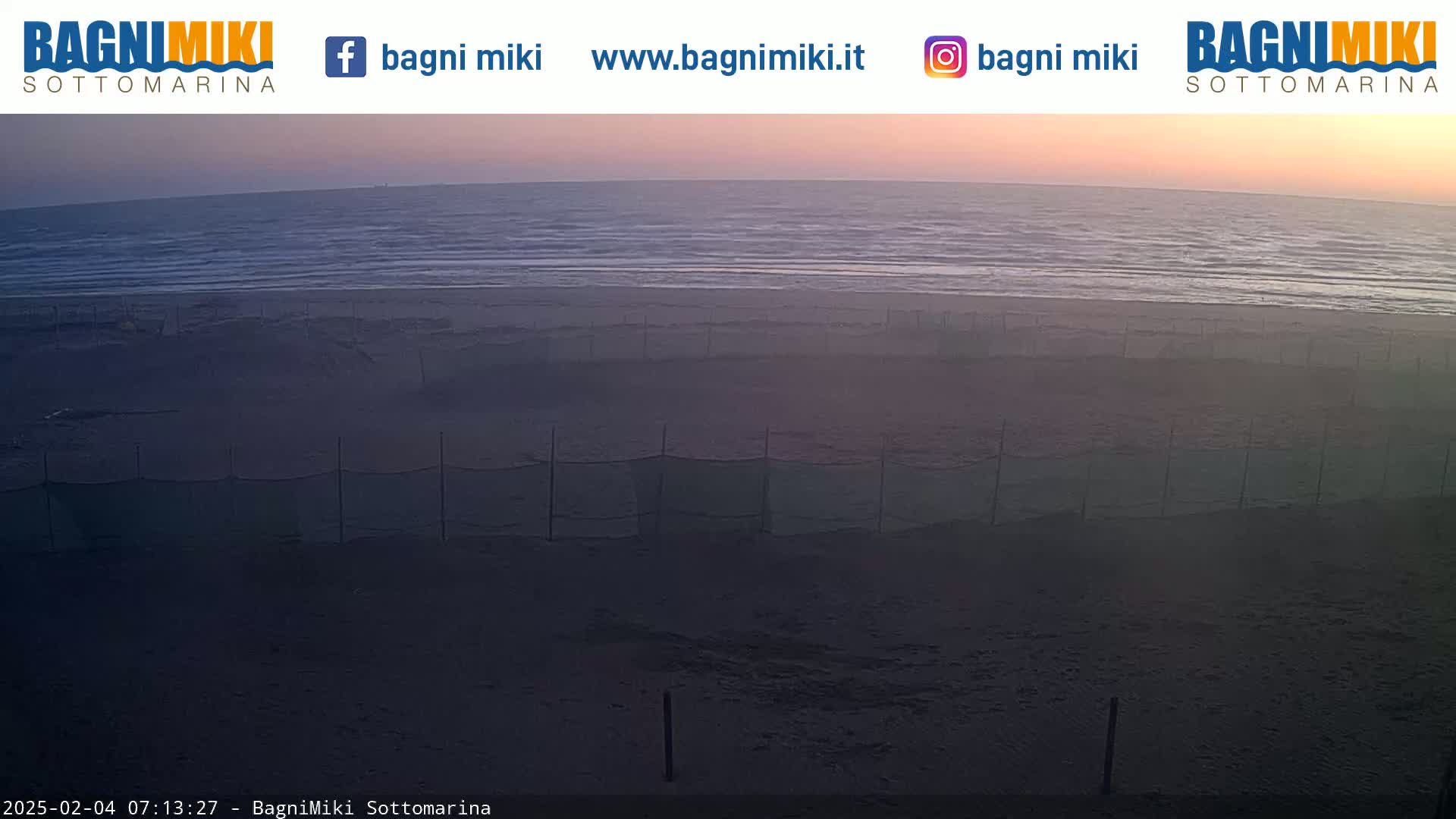 A calm ocean under a dusky pink and orange sunset is visible from a beach with netting separating sections of the sand.