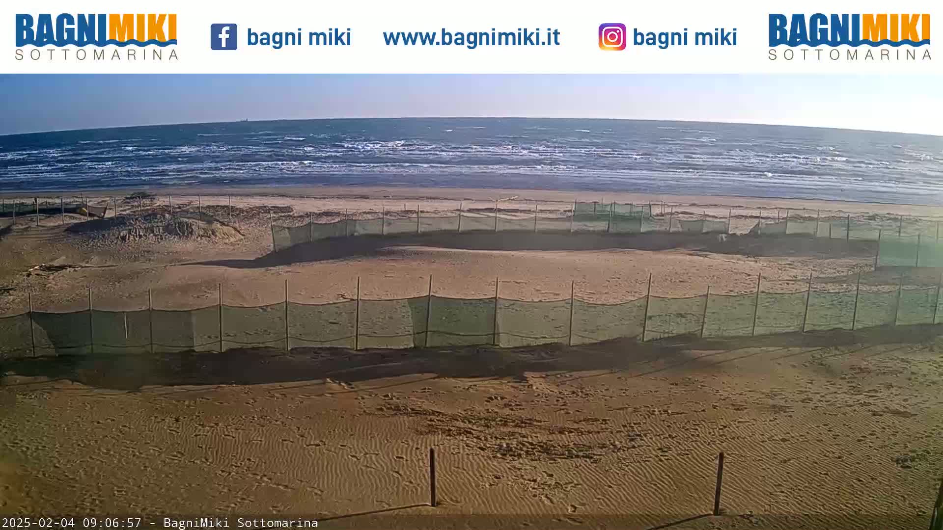 A sandy beach with choppy waves under a clear sky is partially sectioned off by green netting fences.
