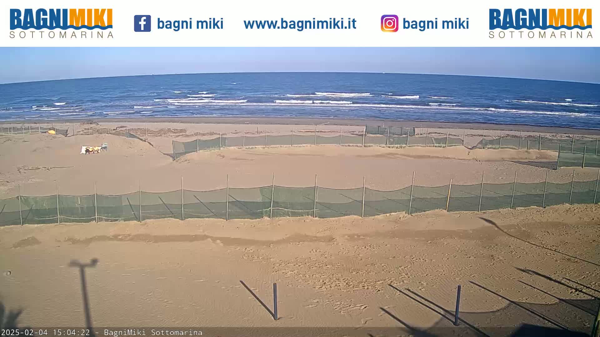 A sandy beach with several fenced-off areas is shown, under a clear sky, with a calm ocean in the background.