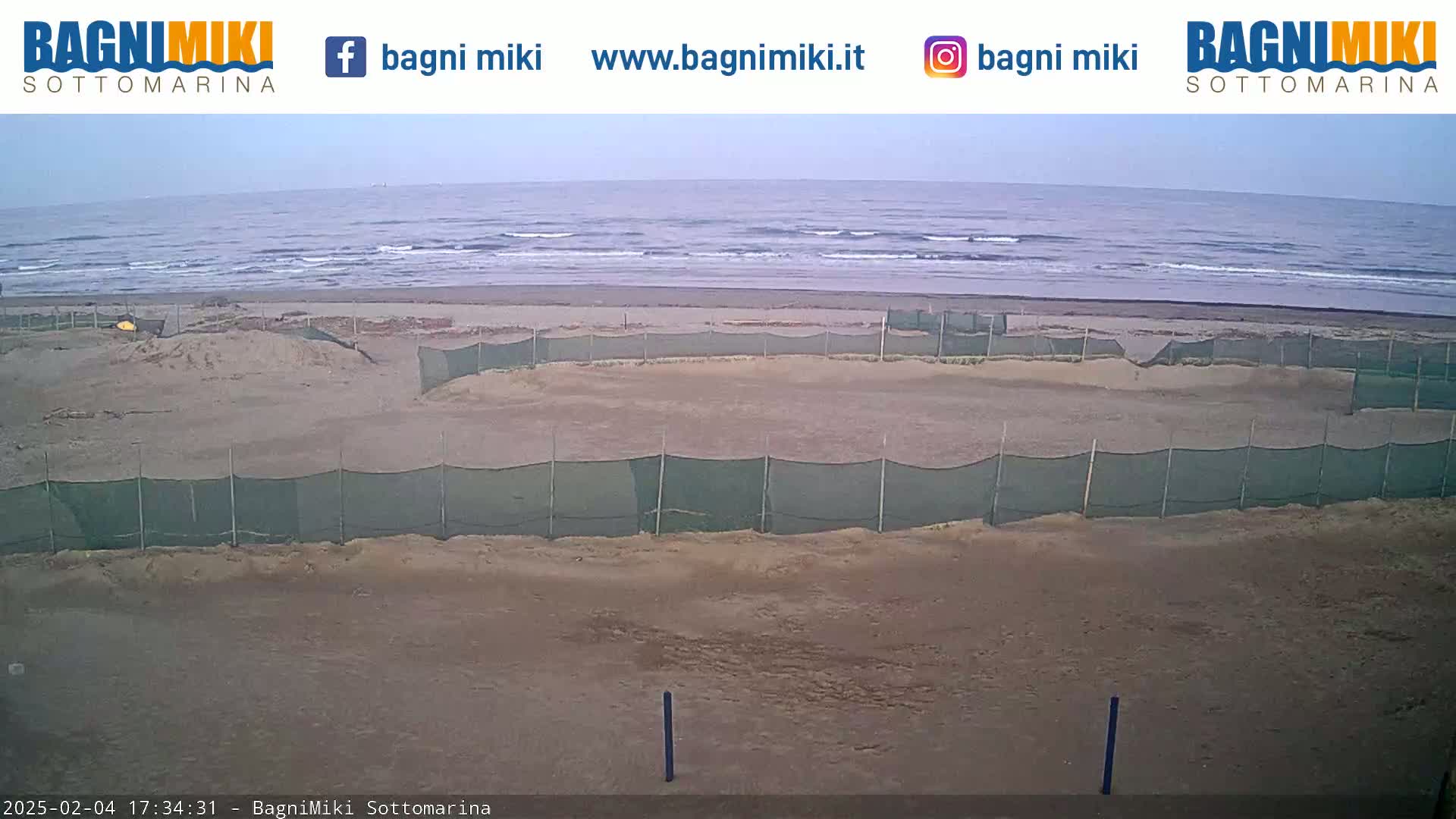 A sandy beach with several enclosed areas sectioned off by netting, overlooks a calm ocean under an overcast sky.