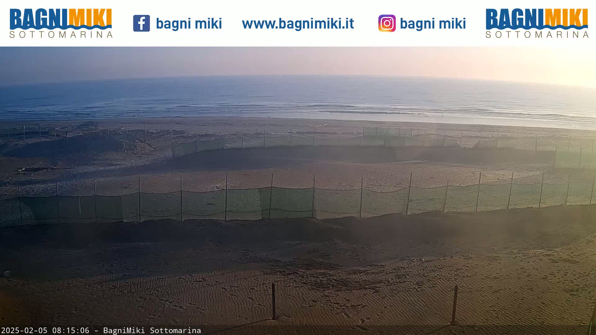 A sandy beach with calm ocean waves under a hazy, sunlit sky is sectioned off by several short green fences.