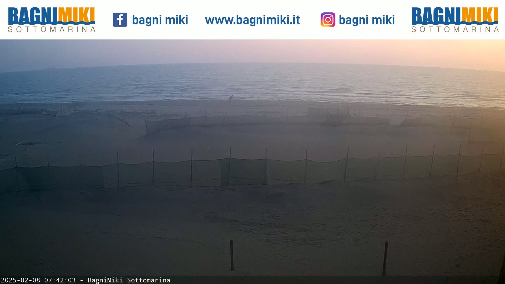 A calm, dusky-colored sea meets a sandy beach protected by several mesh fences under a hazy sky.