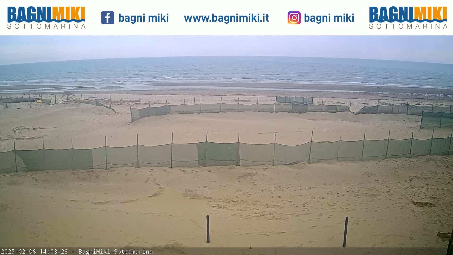 A sandy beach with a calm sea under an overcast sky is enclosed by several sections of netting.