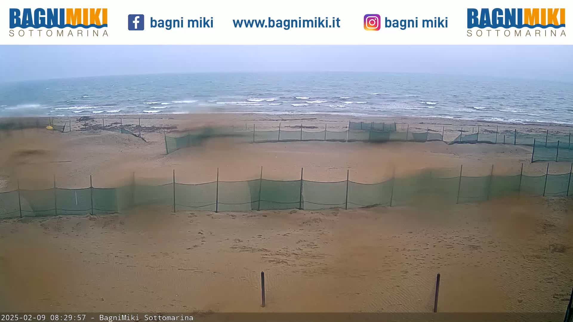 A sandy beach with several sections enclosed by green netting is visible under an overcast sky, with a calm sea in the background.