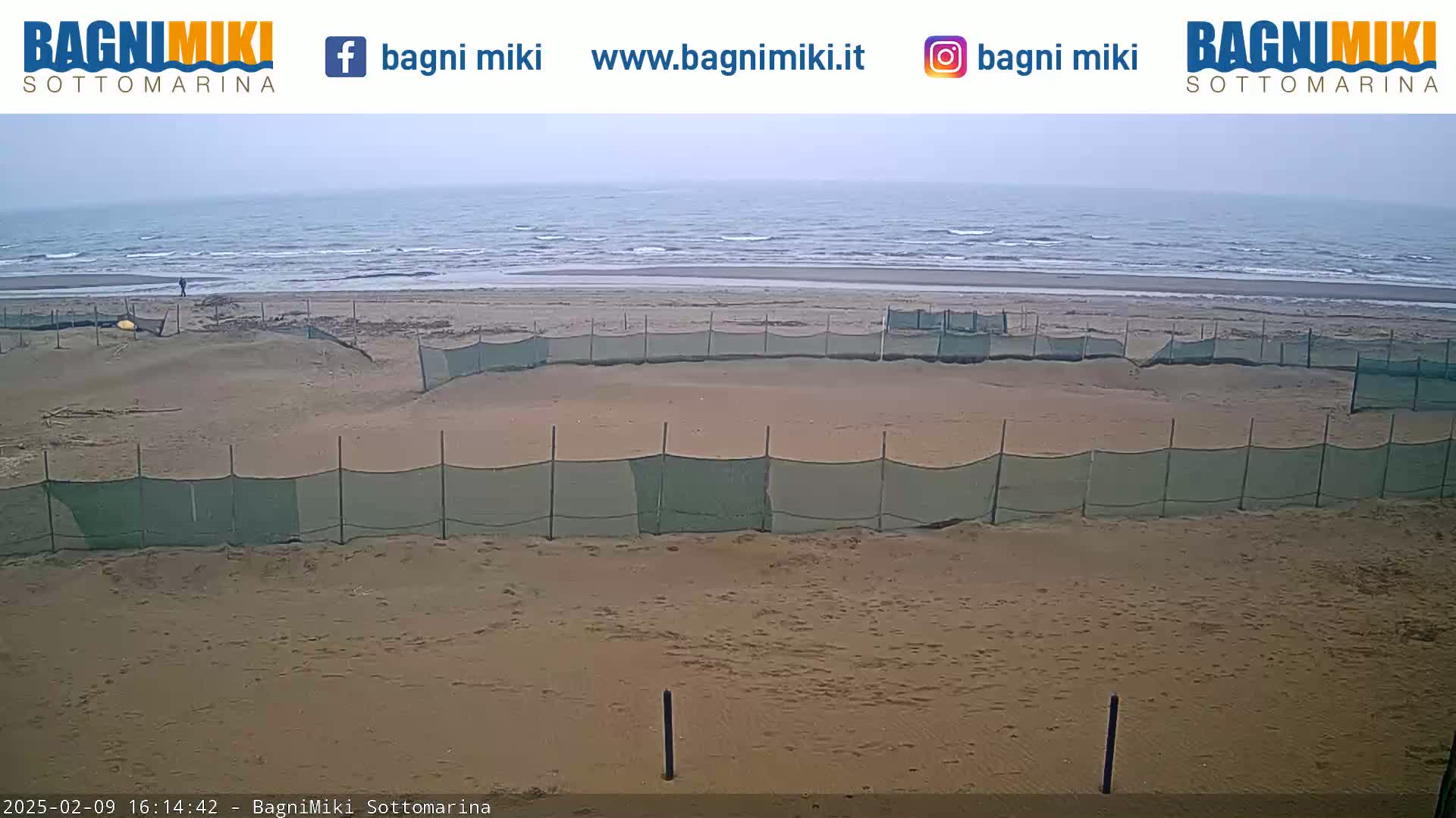 A sandy beach with several sections enclosed by netting is visible, with a calm, overcast sea in the background.