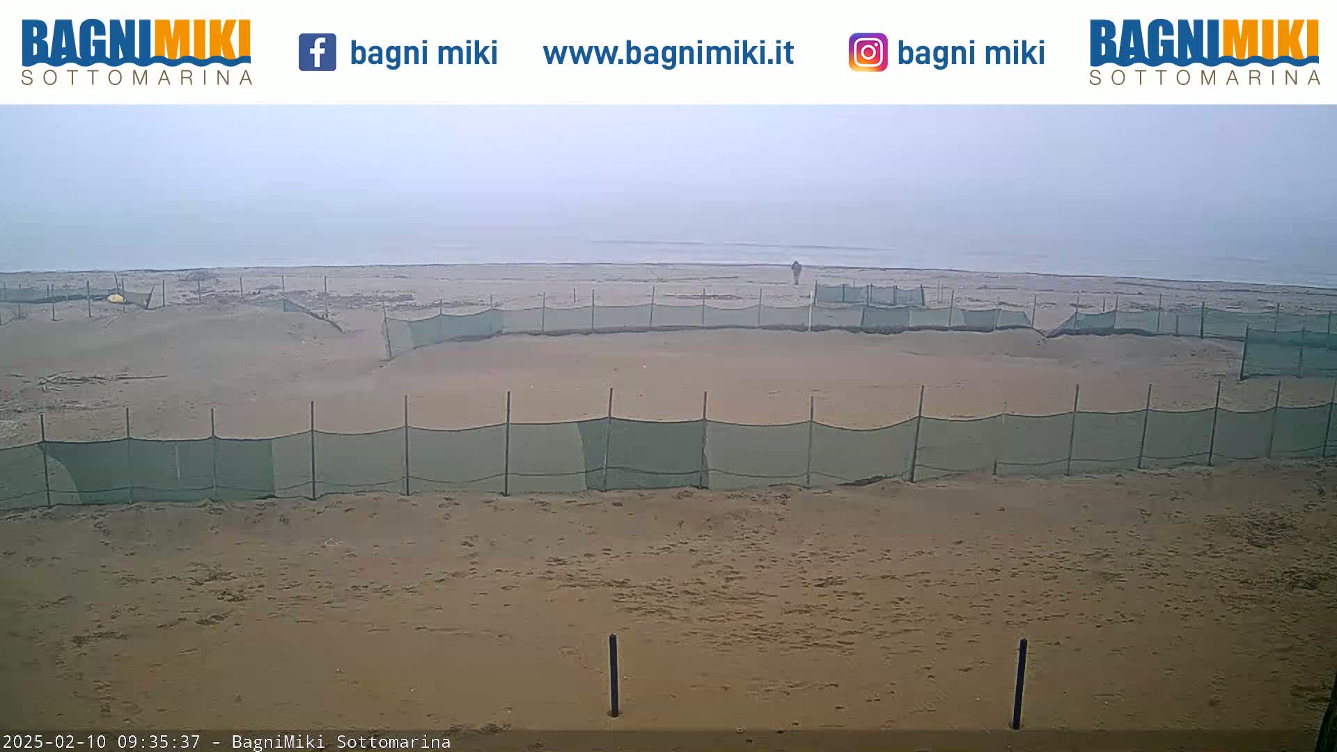 A hazy beach scene shows a sandy shore with several sections enclosed by low green netting, and a lone person walking near the water's edge.