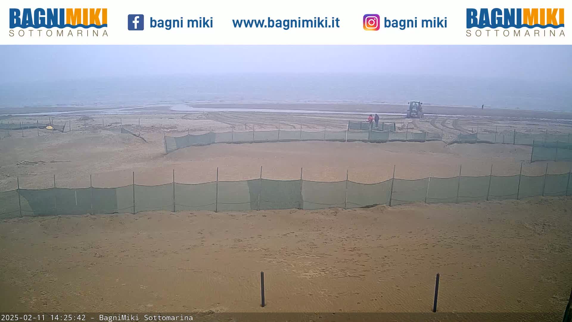 A sandy beach under an overcast sky is partially enclosed by a mesh fence, with a tractor and a few people visible in the background.
