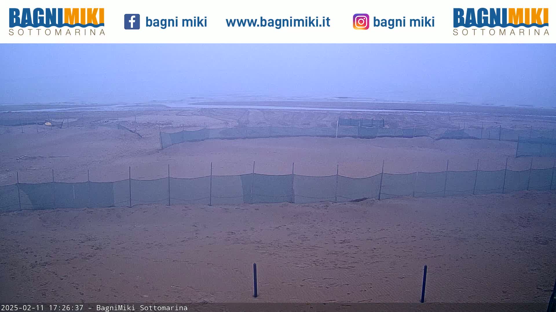 A hazy beach scene shows a sandy shoreline with several mesh fences sectioning off portions of the beach under an overcast sky.