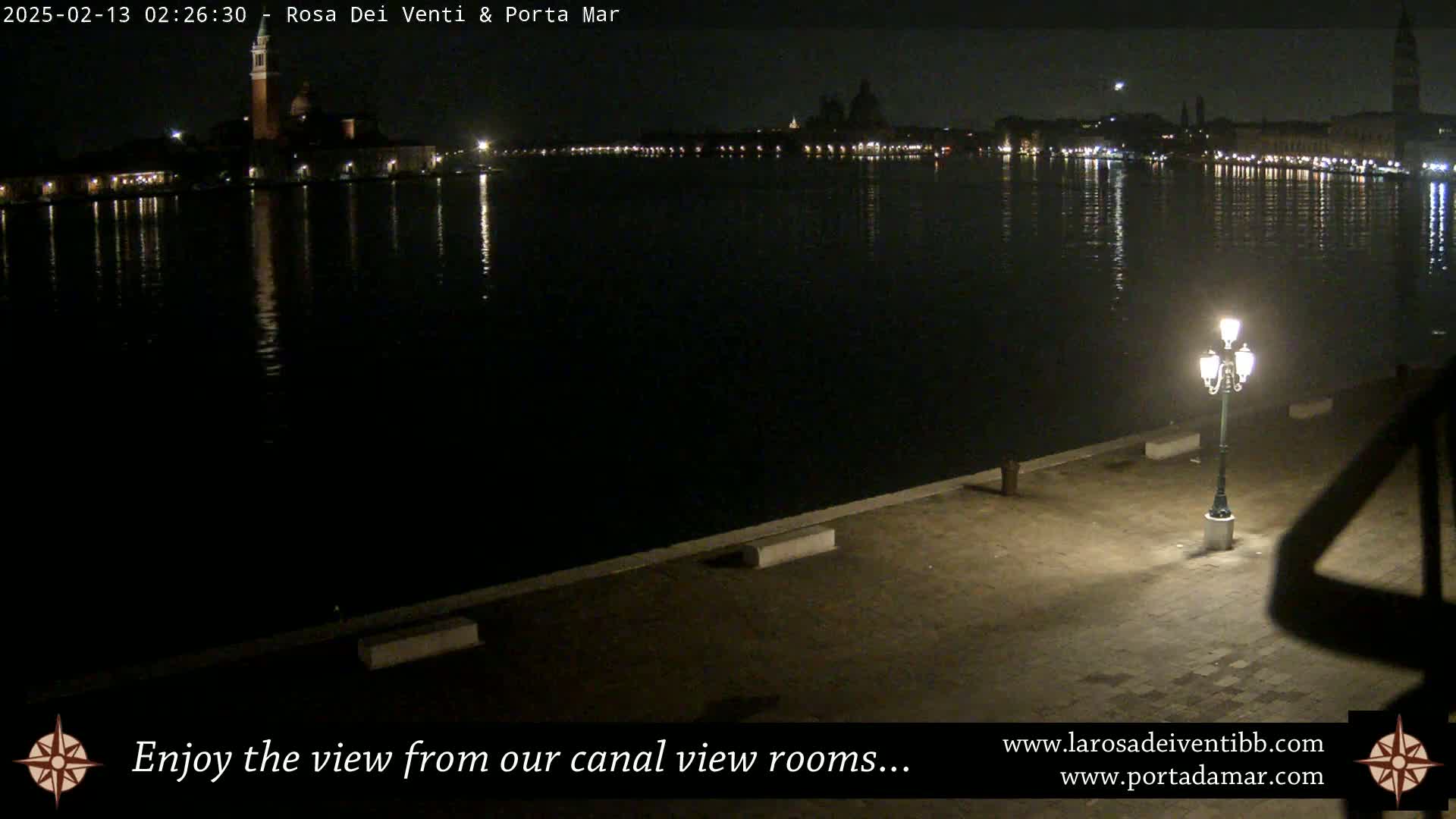 A nighttime view across a calm body of water shows the lights of a distant city reflecting on the water's surface, illuminated by a single lamppost in the foreground.