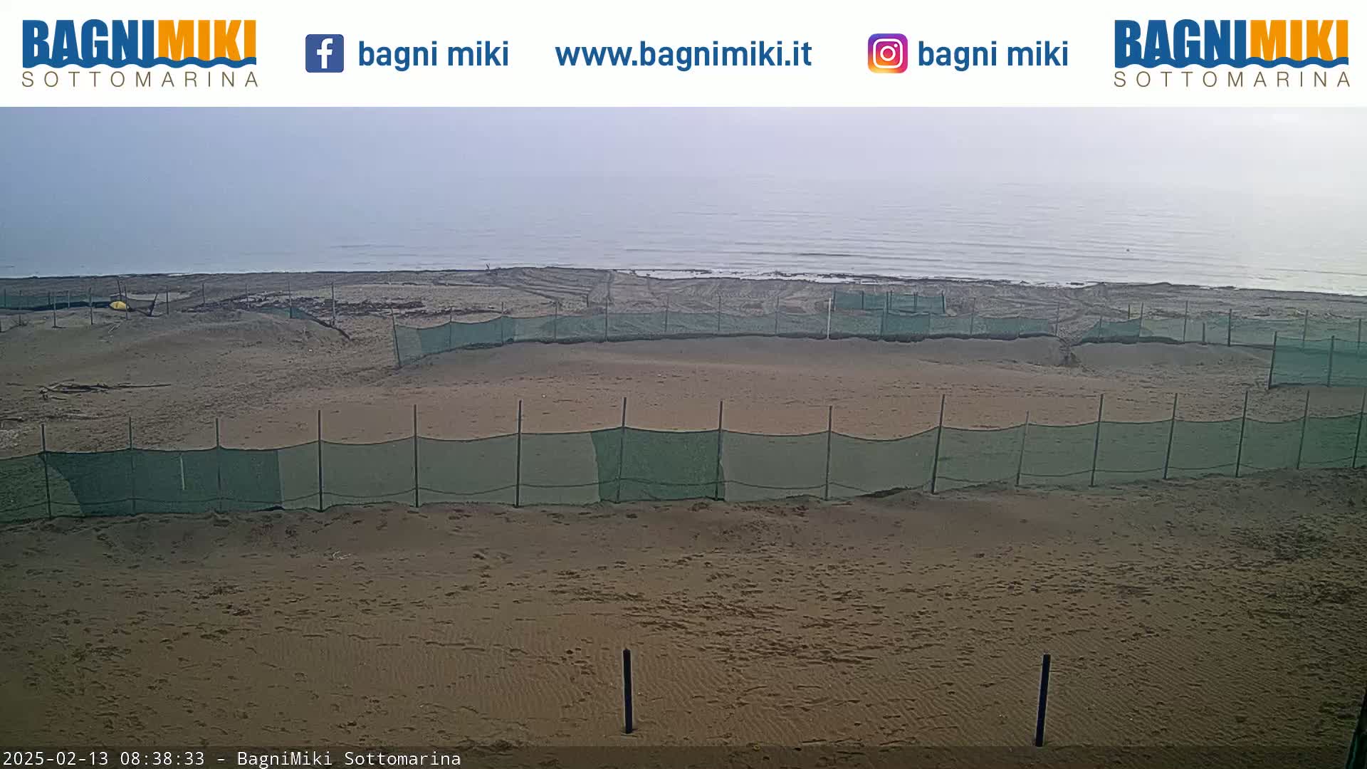 A sandy beach with several green mesh fences is visible, under an overcast sky, next to a calm body of water.