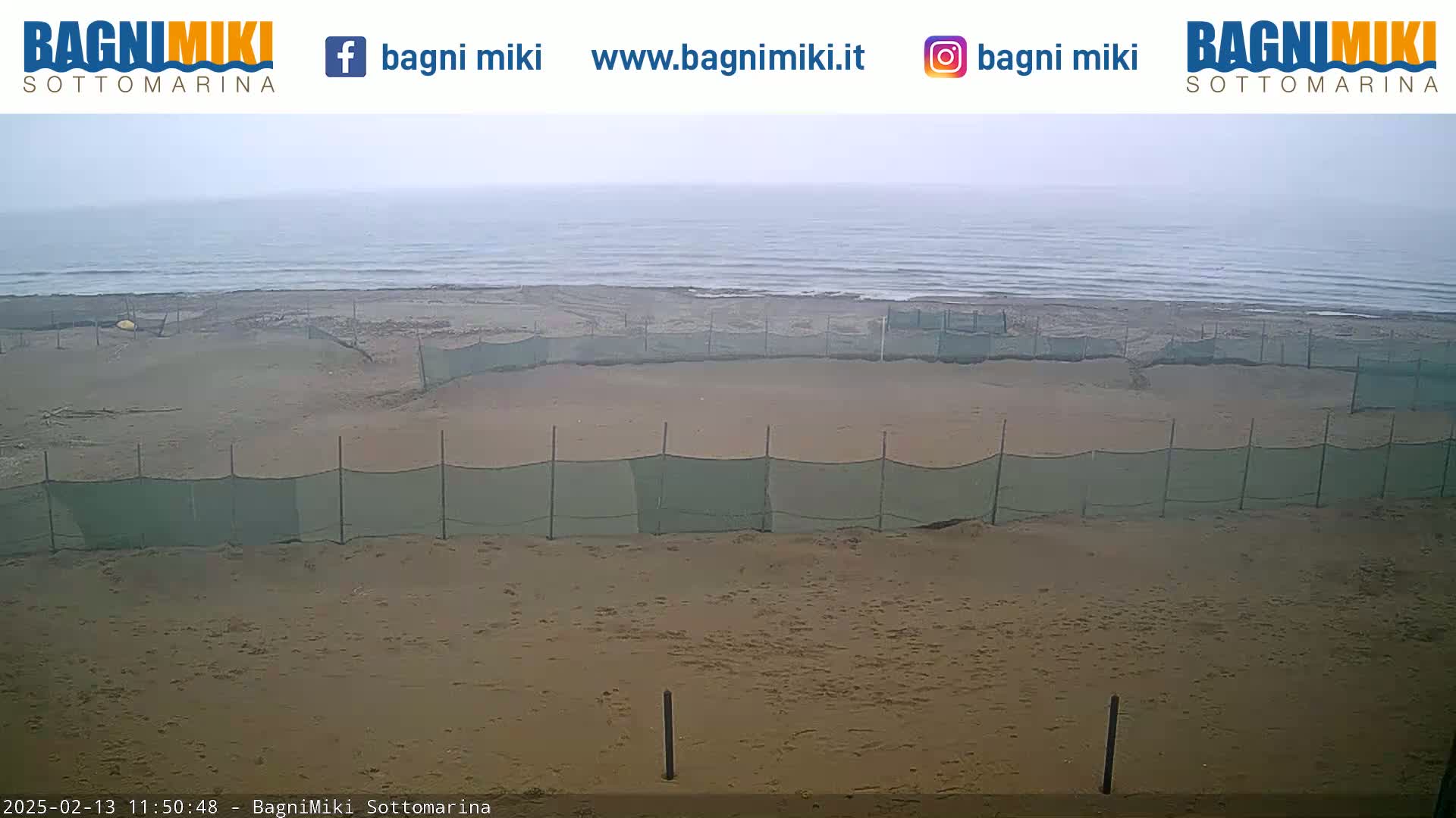 A sandy beach with a calm, overcast ocean in the background is partially sectioned off by green mesh fencing.