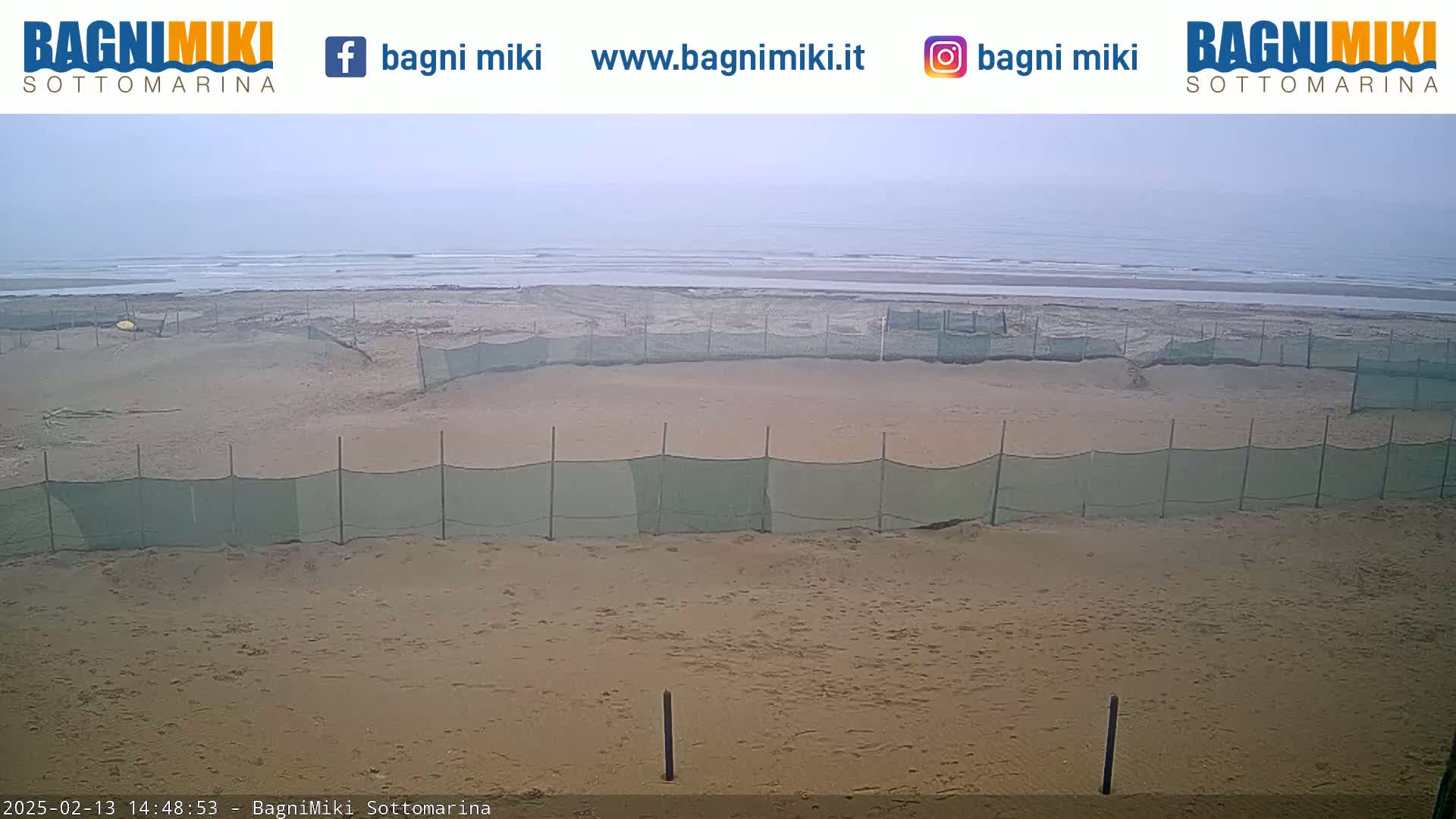 A sandy beach with several sections enclosed by green netting is visible, overlooking a calm, overcast ocean.