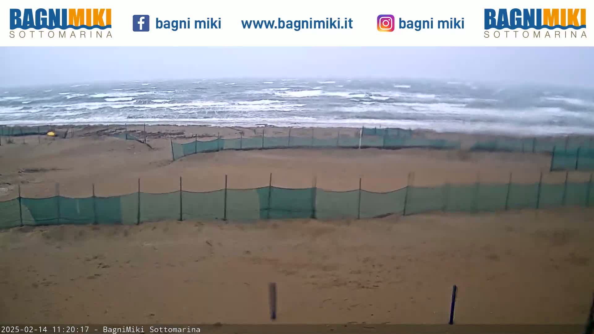 A sandy beach with green netting fences is shown, with choppy waves and an overcast sky visible in the background.