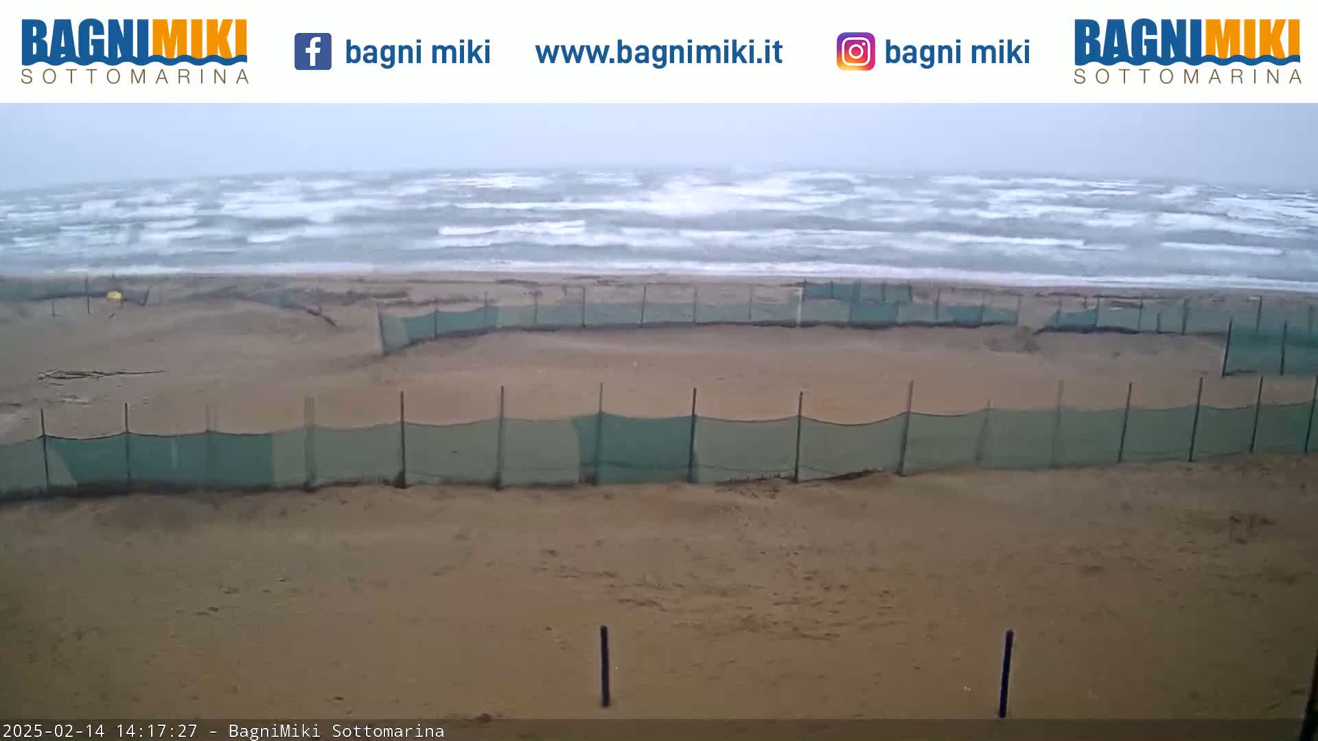 A sandy beach with choppy waves and green mesh fencing is visible under overcast skies.