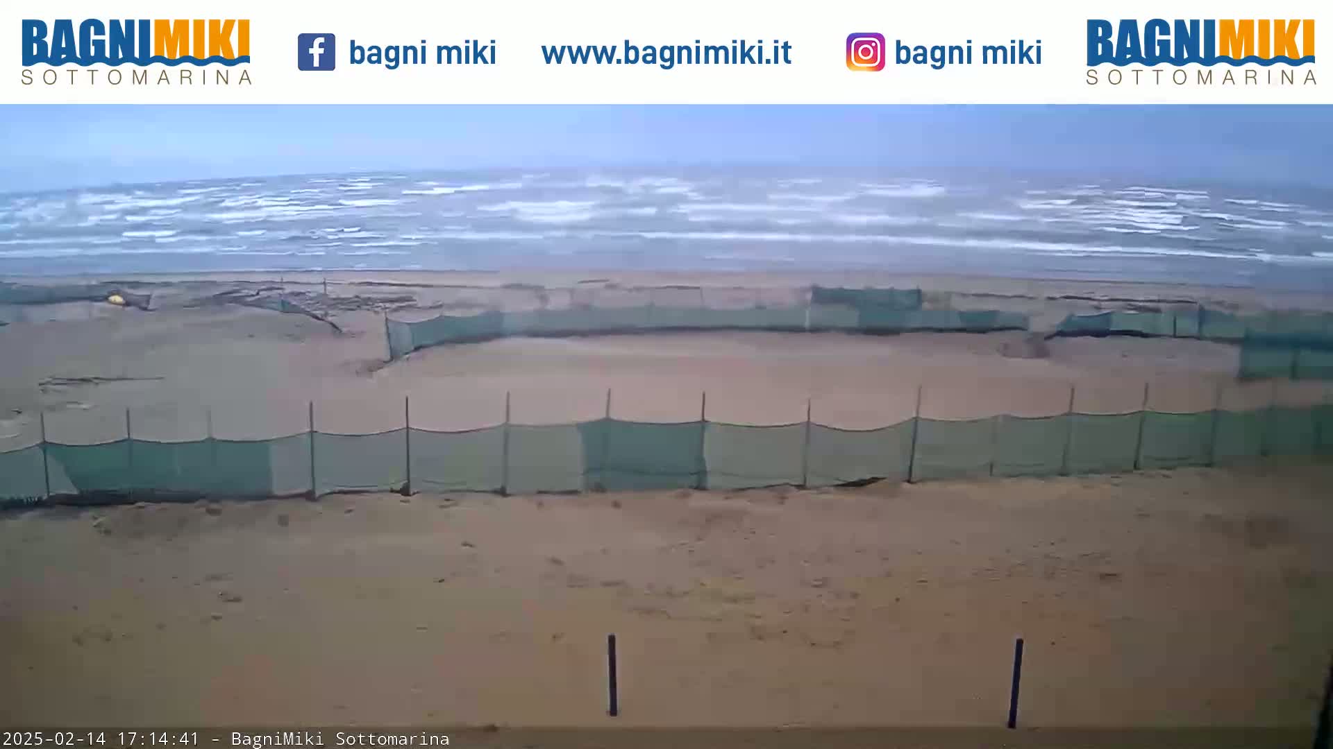 A sandy beach with a section enclosed by a green mesh fence is visible, with a somewhat overcast sky and a wavy ocean in the background.