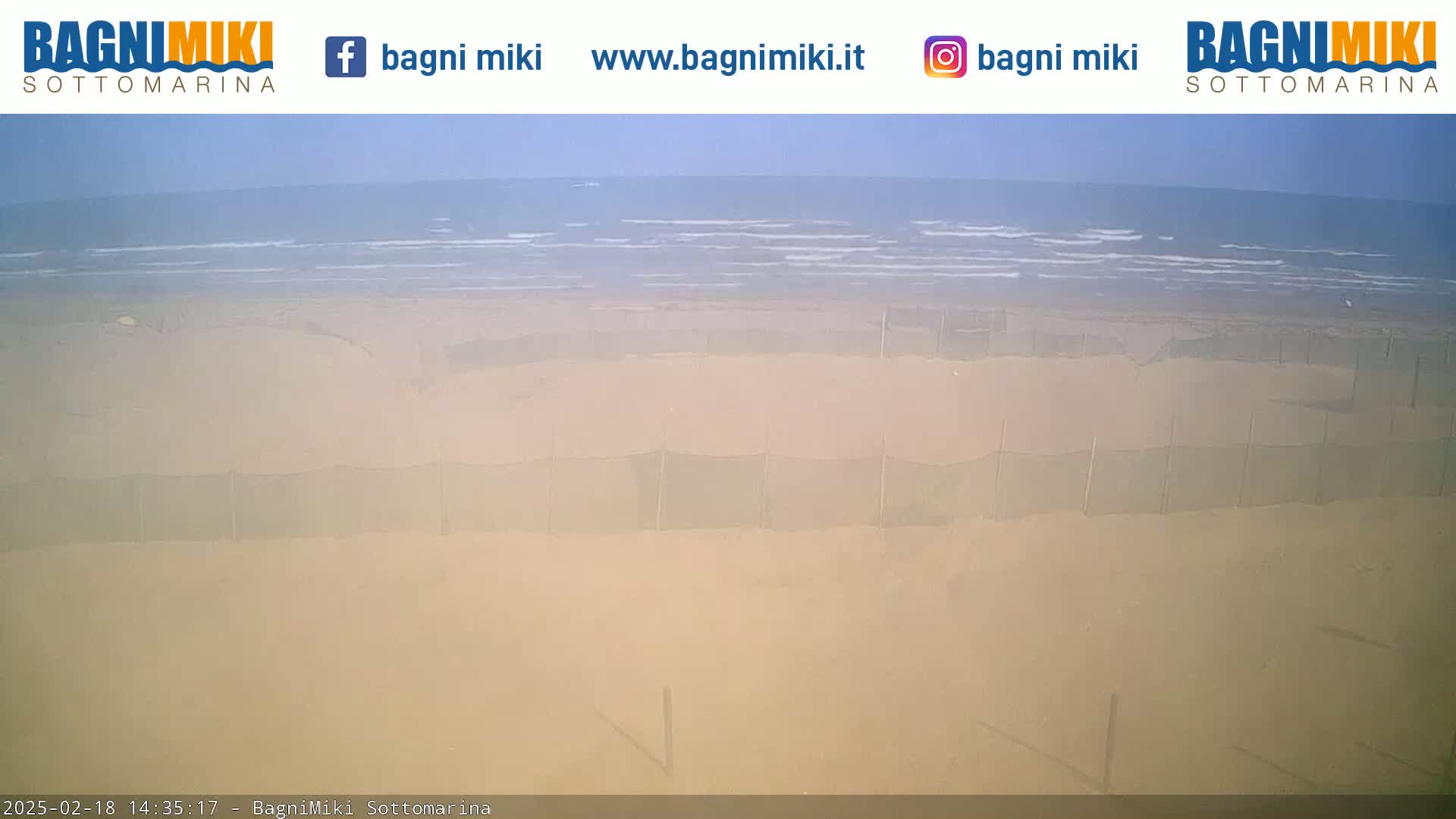 A sandy beach with calm ocean waves and several dark nets or fences is visible under a hazy sky.