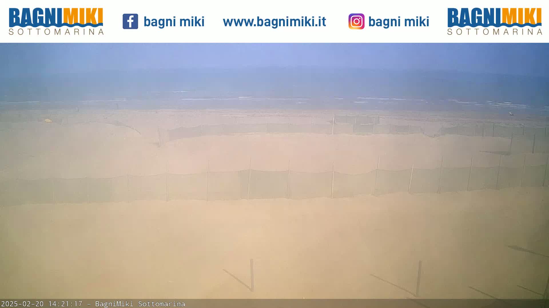 A hazy, sandy beach with several mesh fences stretches to a calm ocean under a light blue sky.