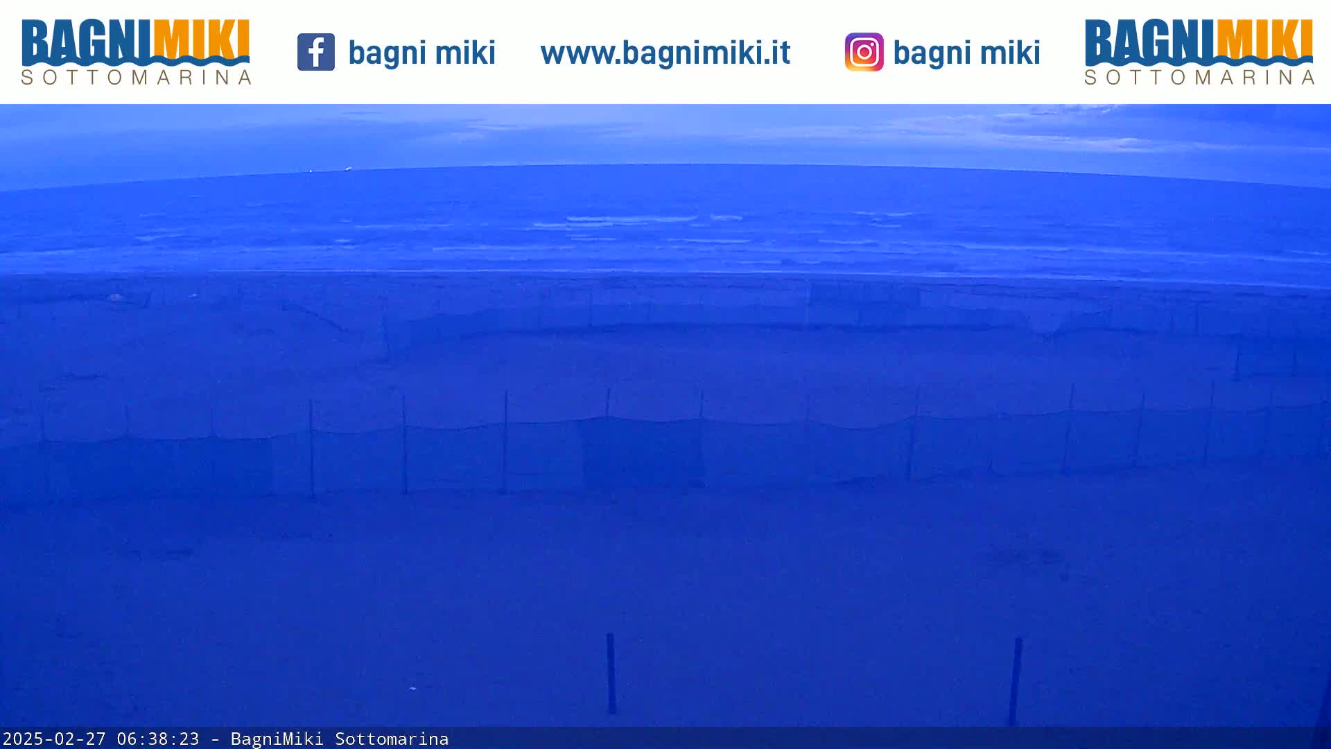 A mostly clear, twilight sky overlooks a dark blue body of water, and a section of what appears to be a fenced-off area on the shore.