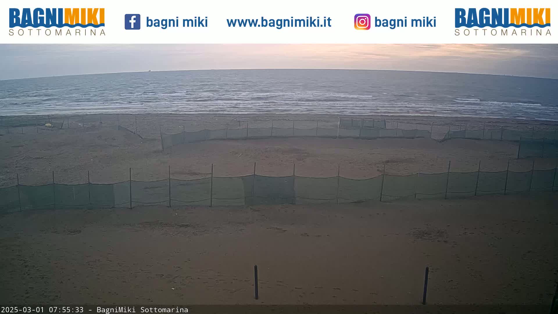 A sandy beach with several sections enclosed by netting is visible, overlooking a calm sea under a soft, dawn-like sky.