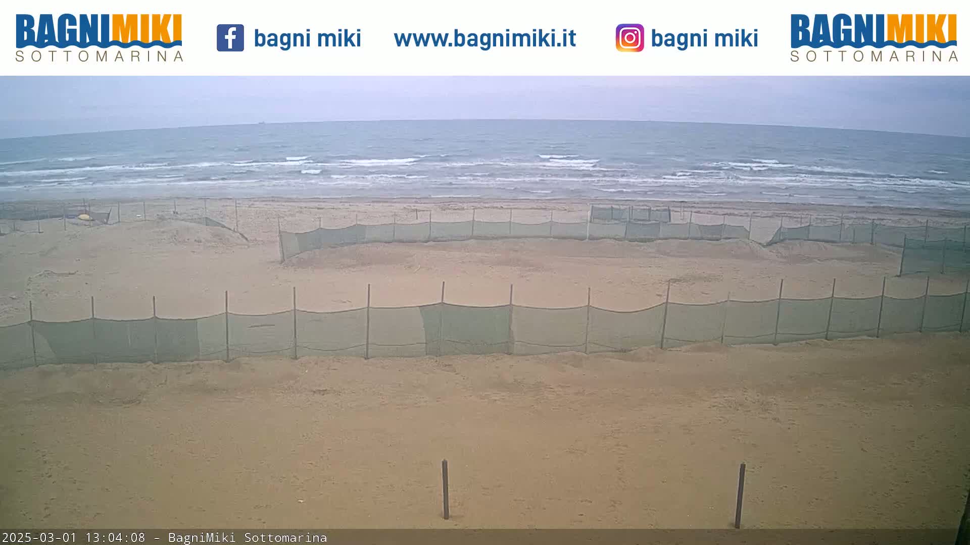 A sandy beach with several enclosed areas marked off by netting is visible, with a somewhat overcast sky and a wavy ocean in the background.