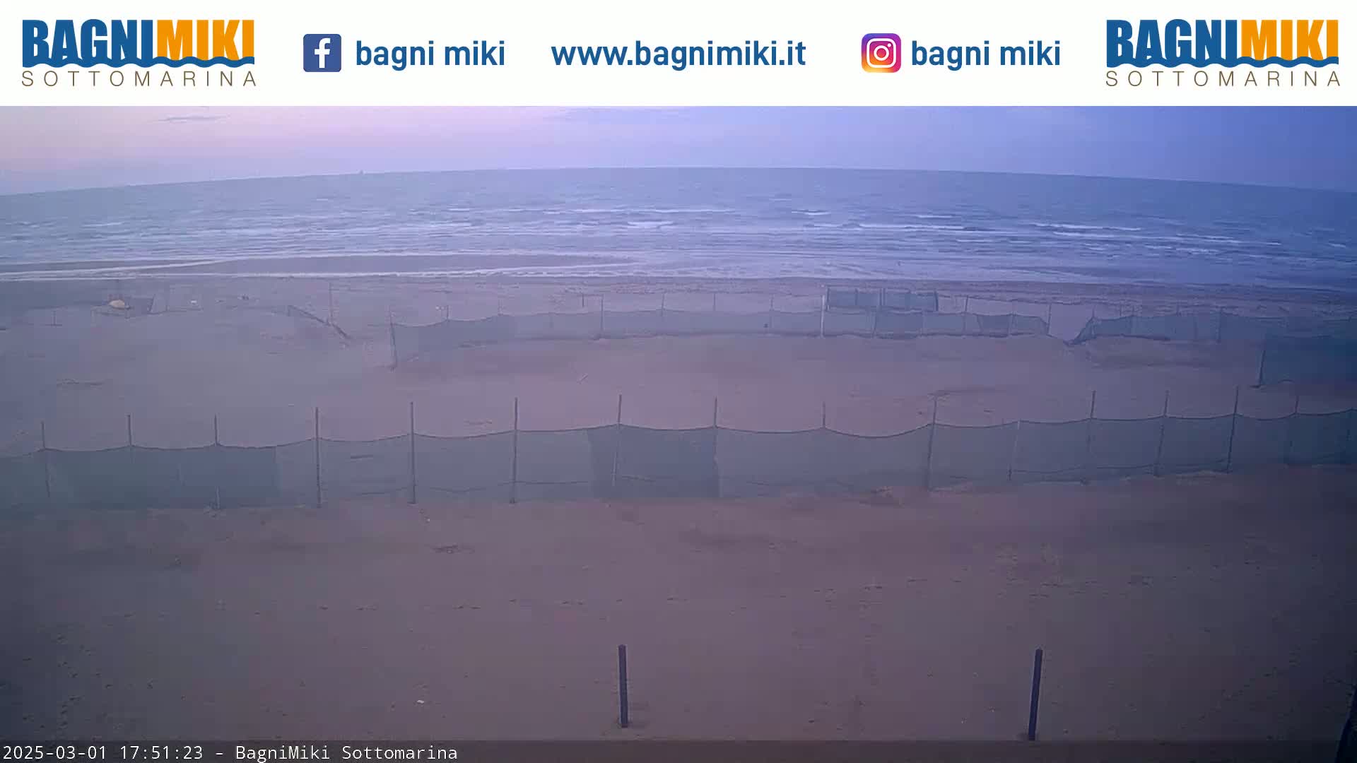 A sandy beach with several mesh fences stretches towards a calm sea under an overcast sky.