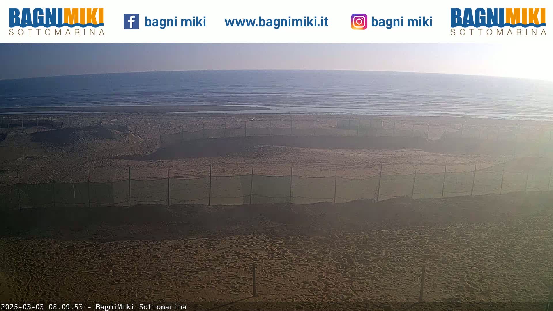 A sandy beach with several sections enclosed by netting is visible, overlooking a calm sea under a hazy sky.