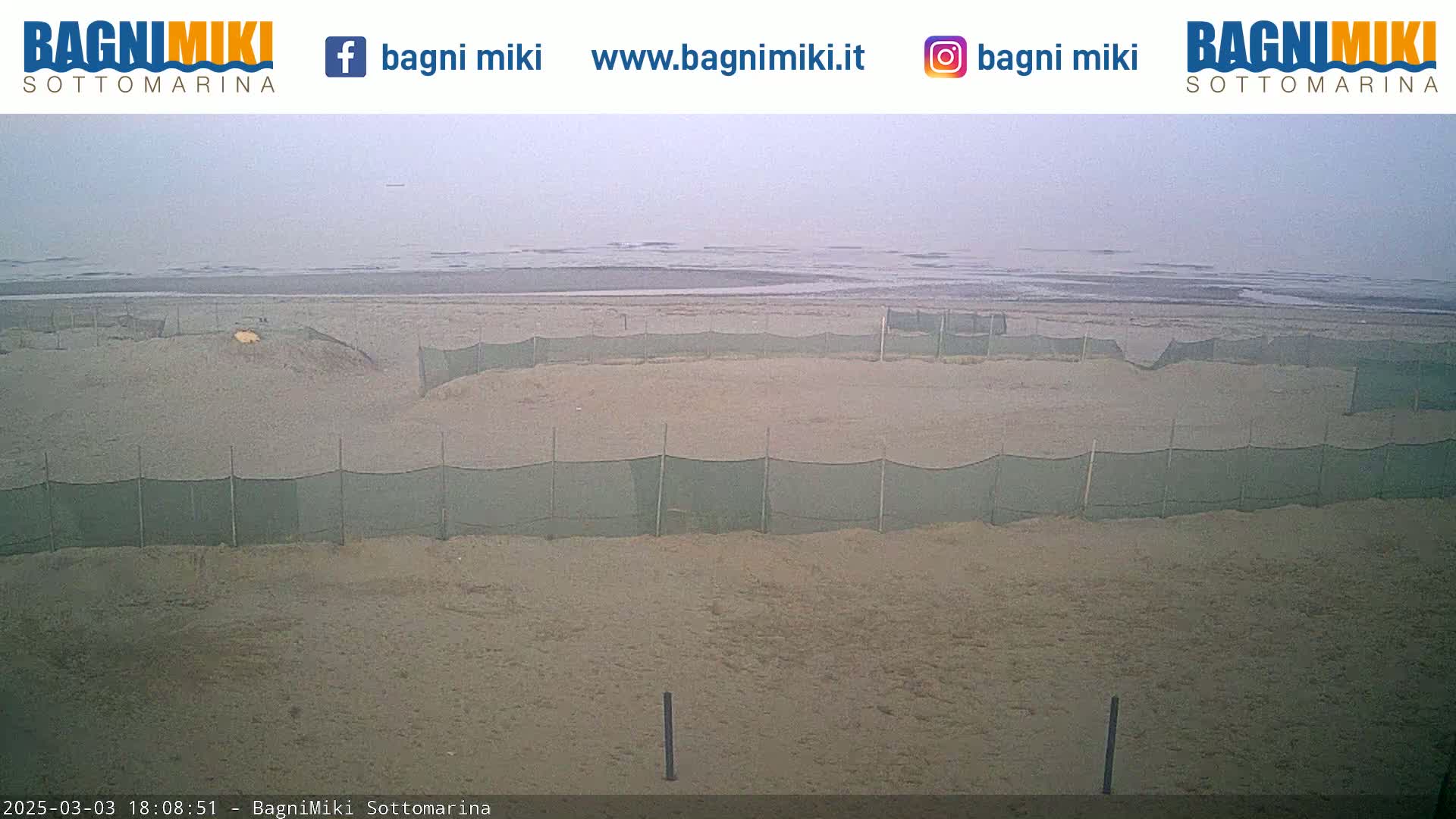 A sandy beach under an overcast sky is partially enclosed by several mesh fences near a calm, gray ocean.