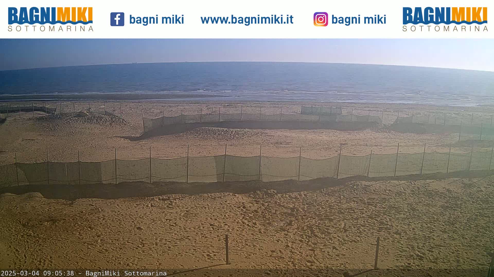 A sandy beach with several areas enclosed by netting is shown, under a bright, sunny sky with a calm ocean in the background.
