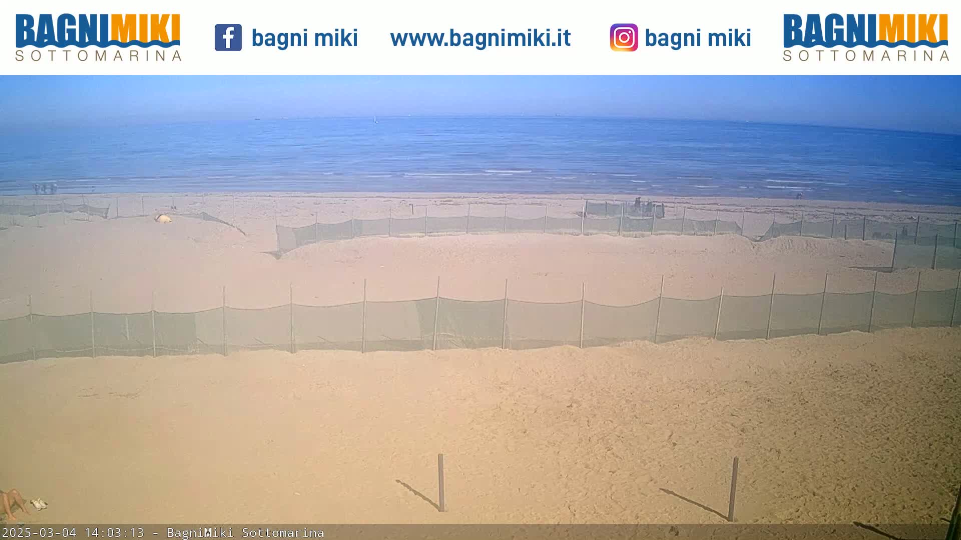A sandy beach with several enclosed areas delineated by mesh fencing is visible, under a clear, sunny sky, with calm ocean water in the background.
