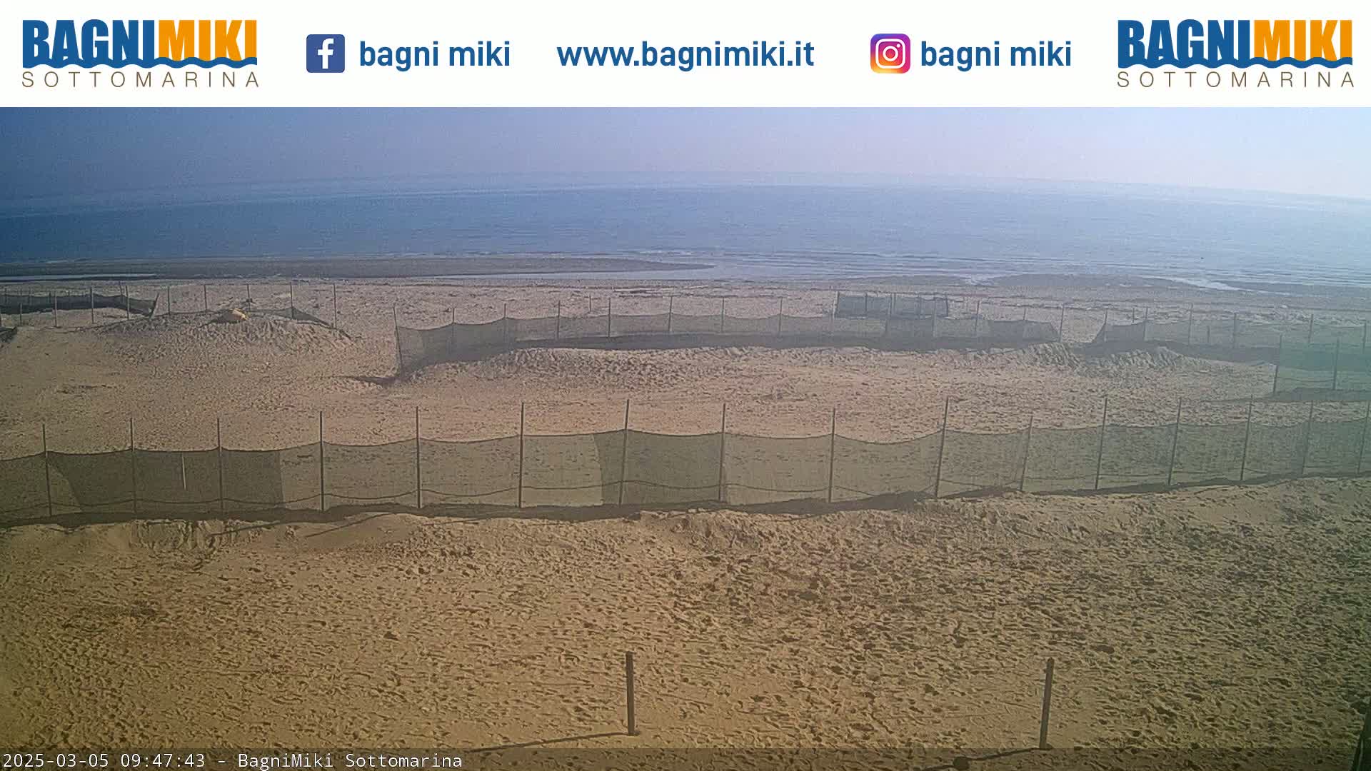 A sandy beach with several fenced-off areas is visible, under a sunny sky, overlooking a calm sea.