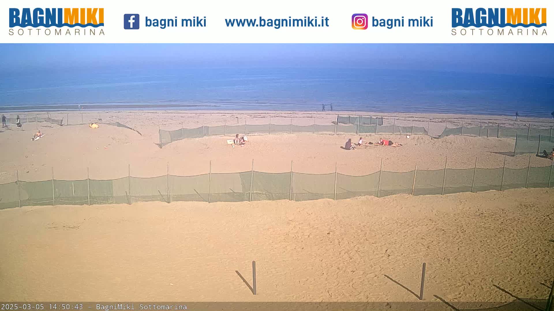 A sandy beach with a calm ocean under a clear sky features several small, enclosed areas on the sand, and a few people are visible.